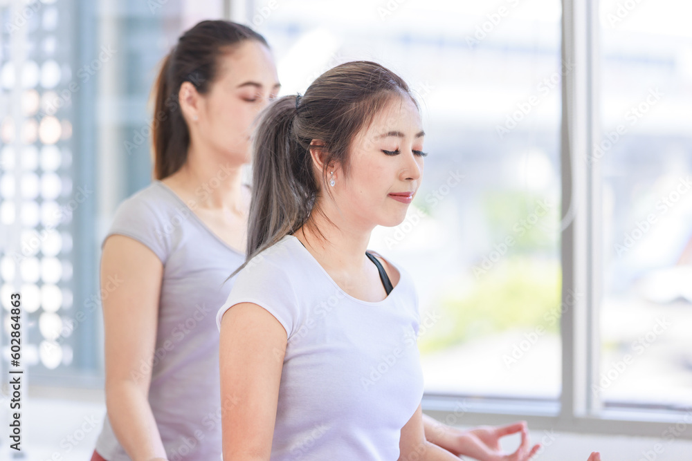 Fototapeta premium Two people Asian beautiful healthy fit slim female sporty athlete model in sport bra and leggings sitting crossed legs in lotus posture on yoga carpet smiling doing meditation in living room at home