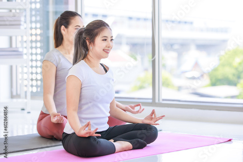 Two people Asian beautiful healthy fit slim female sporty athlete model in sport bra and leggings sitting crossed legs in lotus posture on yoga carpet smiling doing meditation in living room at home