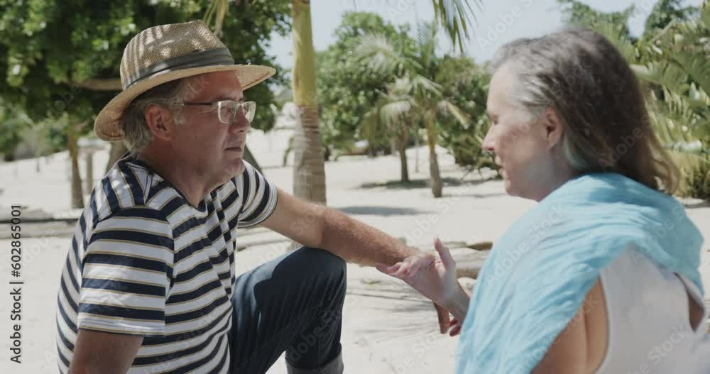 Happy senior caucasian couple sitting on beach talking in the sun, in slow motion
