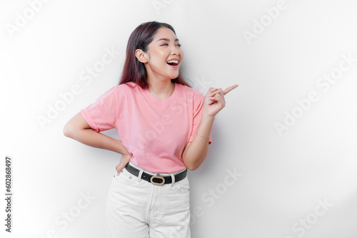Excited Asian woman is wearing pink t-shirt, pointing at the copy space beside her, isolated by white background