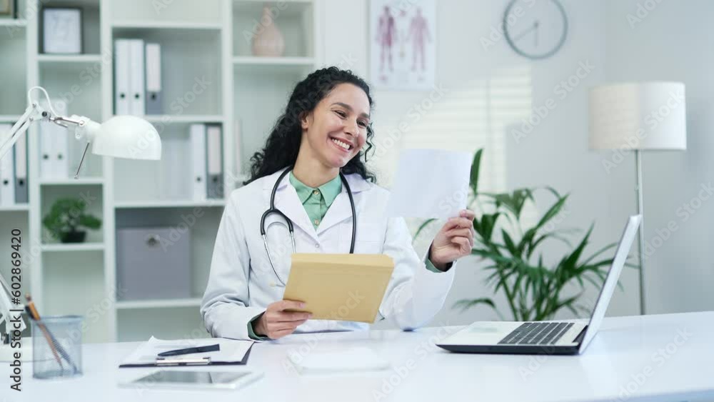 Happy smiling female doctor in white coat received letter with great news sitting in office at workplace in modern hospital clinic. Joyful medical worker physician rejoices at a pleasant notification Happy smiling female doctor in white coat received letter with great news sitting in office at workplace in modern hospital clinic. Joyful medical worker physician rejoices at a pleasant notification