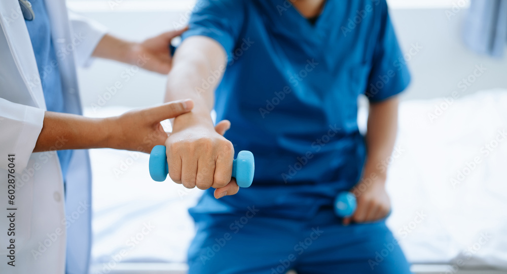 Asian physiotherapist helping male patient stretching arm during exercise correct with dumbbell in hand during training hand in hospital office. Asian physiotherapist helping male patient stretching arm during exercise correct with dumbbell in hand during training hand in hospital office.