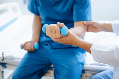 Asian physiotherapist helping male patient stretching arm during exercise correct with dumbbell in hand during training hand in hospital office.