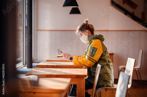 Young blonde woman with ponytail in green yellow raincoat, protective mask on face sitting in cafe