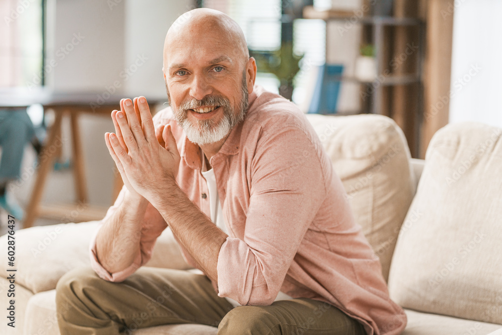 Smiling, happy senior man sitting on a sofa in the comfort of his home. With his hands held together, he radiates a sense of contentment and relaxation. Smiling, happy senior man sitting on a sofa in the comfort of his home. With his hands held together, he radiates a sense of contentment and relaxation.