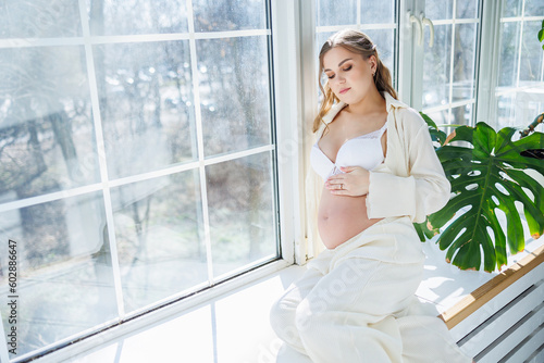 A stylish young pregnant woman in a white suit is sitting on the windowsill. Pregnant woman in white underwear.