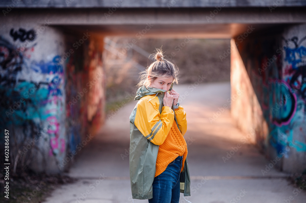 Young blonde girl in yellow-green raincoat with funny ponytail stands on road before graffity tonnel