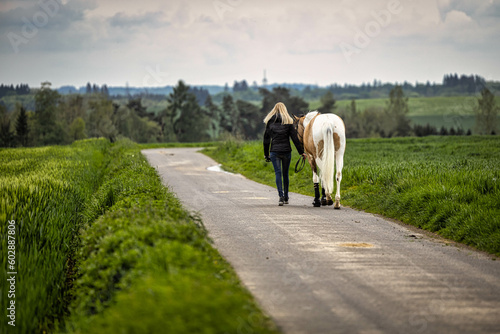Horse with woman go over a paved dirt road, photographed from behind.