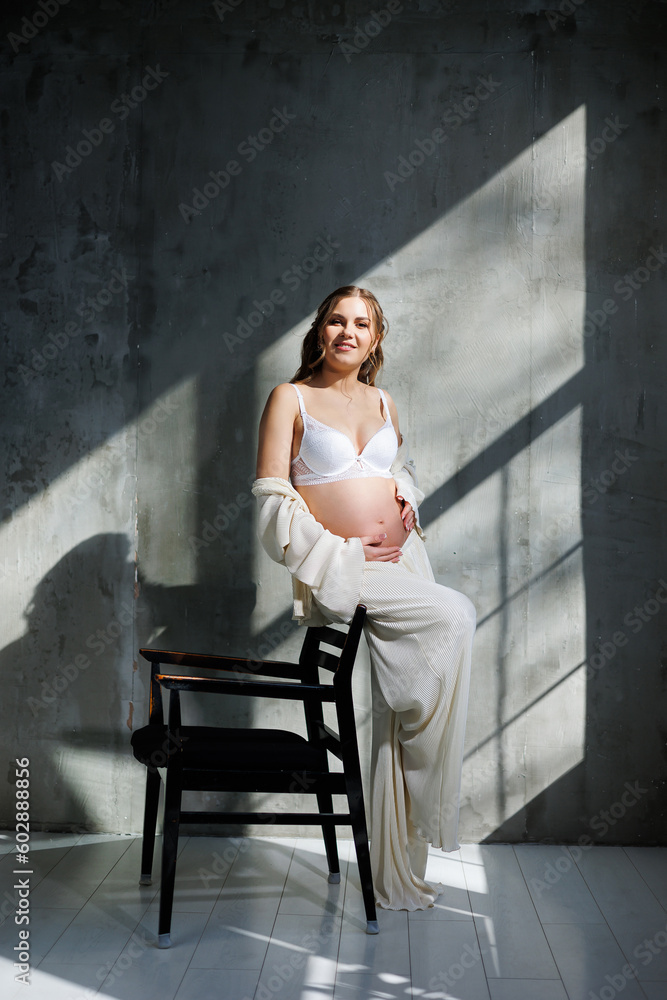 A stylish young pregnant woman in a white suit is sitting on a chair on a gray background. Pregnant woman in white underwear. A stylish young pregnant woman in a white suit is sitting on a chair on a gray background. Pregnant woman in white underwear.