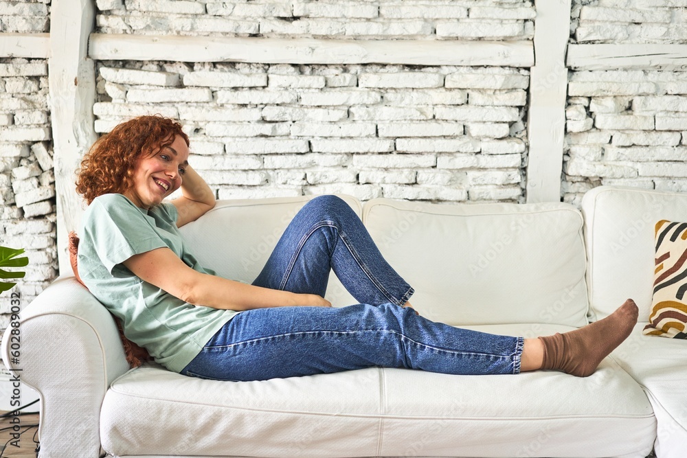 Obraz premium Relaxed young redhead woman lounging on her sofa at home, with a content smile.