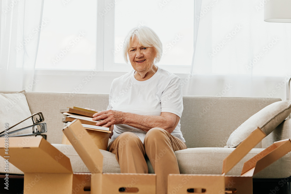 elderly woman sits on a sofa at home with boxes. collecting things with memories albums with photos and photo frames moving to a new place cleaning things and a happy smile. Lifestyle retirement. elderly woman sits on a sofa at home with boxes. collecting things with memories albums with photos and photo frames moving to a new place cleaning things and a happy smile. Lifestyle retirement.