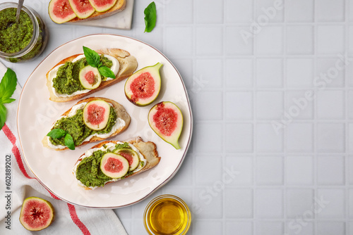 Tasty bruschettas with cream cheese, pesto sauce, figs and fresh basil on white tiled table, flat lay. Space for text