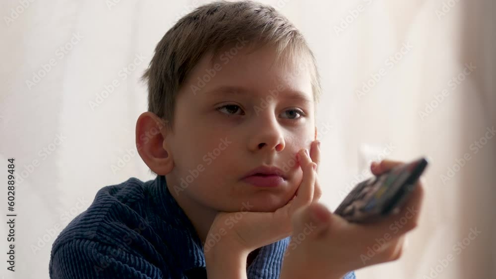 portrait of a handsome european boy watching tv holding a tv remote in his hands while being indoors.lifestyle of a schoolboy at home. bored child watching movie portrait of a handsome european boy watching tv holding a tv remote in his hands while being indoors.lifestyle of a schoolboy at home. bored child watching movie