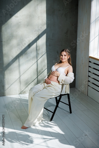 Stylish pregnant young woman in a white suit near the window and touching her belly with a gentle smile. Awaiting birth. Care and motherhood. Love and tenderness.