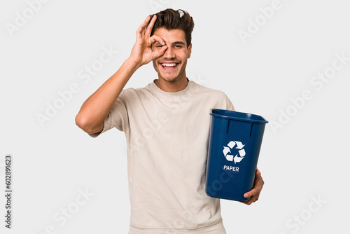 Young caucasian man holding a paper trash for recycle excited keeping ok gesture on eye.