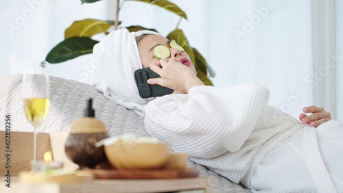 Woman laying on spa bed feeling relax with cucumber slices on eyes and talking by smartphone at spa room. Spa aromatherapy concept