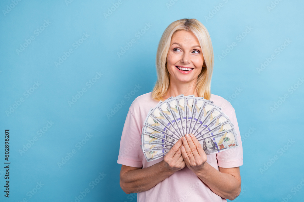 Portrait of successful senior woman with bob hairstyle wear white t-shirt look empty space hold dollars isolated on blue color background