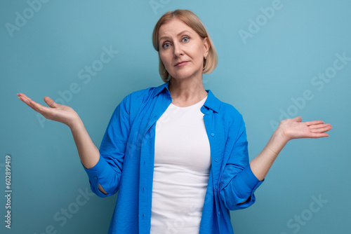 blond mature woman throws up her hands in ignorance on a blue background with copyspace