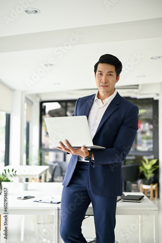 Successful Asian businessman in formal business suit stands in his office with his laptop