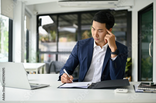 Thoughtful millennial Asian businessman rechecking business documents at his desk. photo