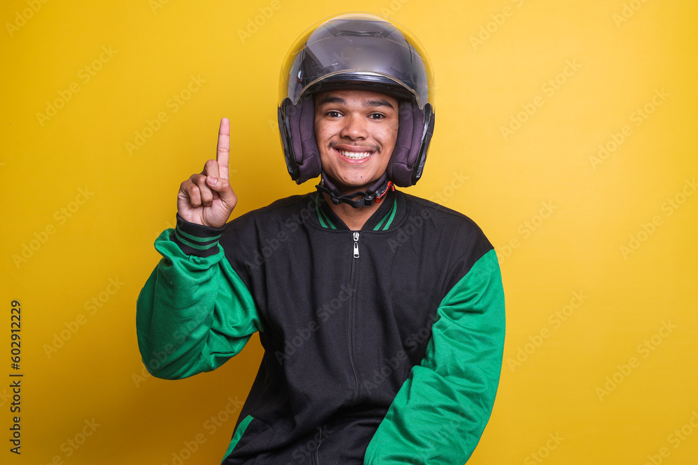 Asian online taxi driver wearing green jacket and helmet pointing to empty space with finger isolated over yellow background Asian online taxi driver wearing green jacket and helmet pointing to empty space with finger isolated over yellow background