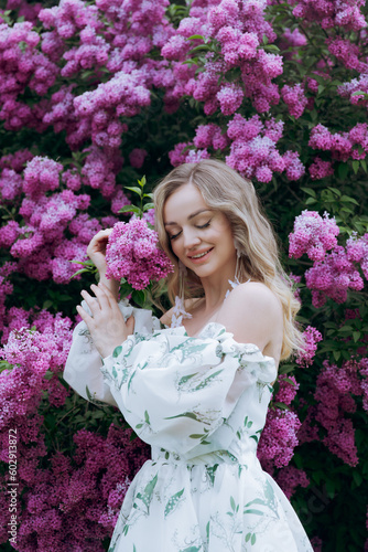 Beautiful, stylish, cute blonde in a white dress with a green pattern as a model for a photo shoot in lilac. They made luxurious portraits of her in the green garden of the park.