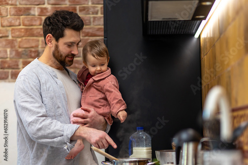 Bearded man cooking dinner with baby daughter in hands standing in kitchenn.