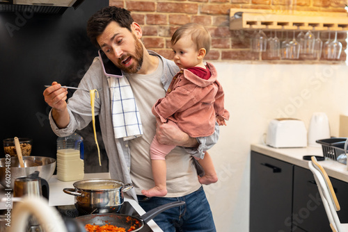 Multitasking father babysitting, cooking breakfast and talking on smart phone at home. photo