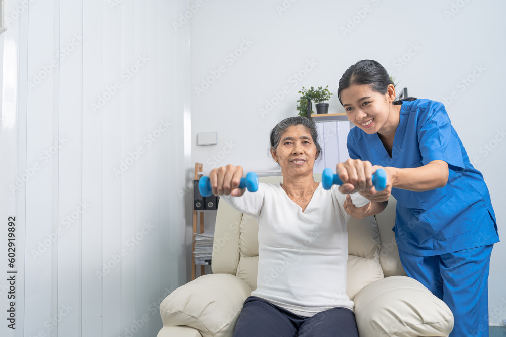 Fototapeta premium Young pretty asian physiotherapist helping senior female patient holding dumbbell in physical therapy session, concept of World Confederation for Physical Therapy.