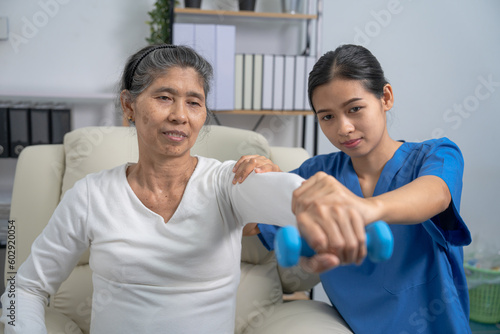 Young pretty asian physiotherapist helping senior female patient holding dumbbell in physical therapy session, concept of World Confederation for Physical Therapy.