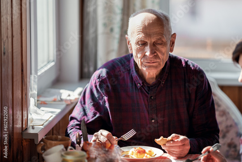 Elderly man eats while sitting at family table in cottage kitchen. Caring for old people concept.