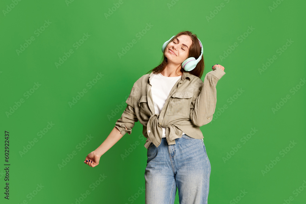 Close up portrait of beautiful young girl, teenager in headphones and listening to music with pleasure over green color studio background Close up portrait of beautiful young girl, teenager in headphones and listening to music with pleasure over green color studio background