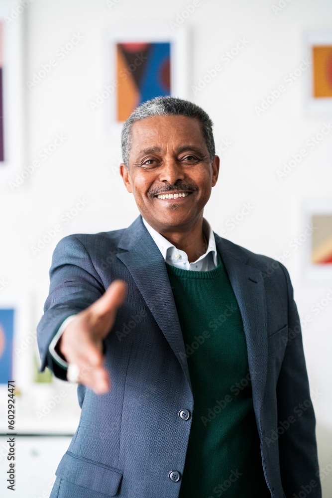 Positive successful mature black entrepreneur in suit smiling while stretching hand at camera in greeting gesture Positive successful mature black entrepreneur in suit smiling while stretching hand at camera in greeting gesture