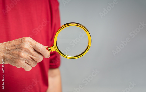 Senior woman wearing a red casual holding a magnifying glass while standing against a gray background
