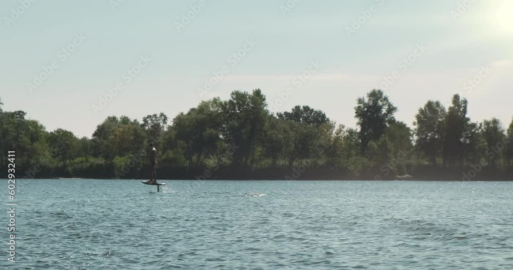 Man standing by electric hydrofoil, summer backdrop. Beautiful river landscape. Active lifestyle on vacation recreation. Modern water sport. Lift Foil Electric Hydrofoil Surfboard. Weekend near river Man standing by electric hydrofoil, summer backdrop. Beautiful river landscape. Active lifestyle on vacation recreation. Modern water sport. Lift Foil Electric Hydrofoil Surfboard. Weekend near river
