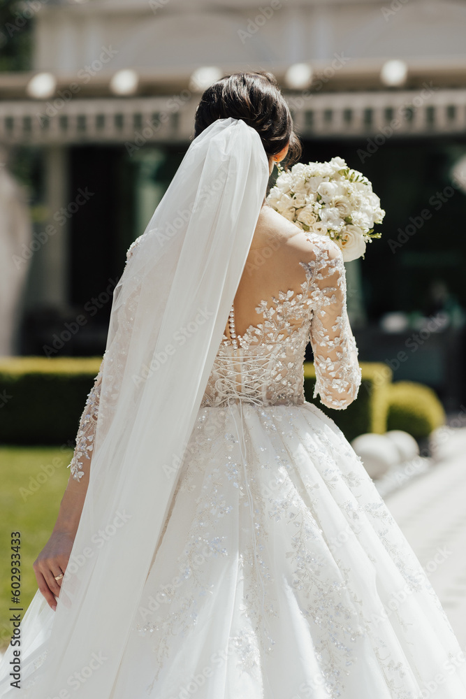 Back view of young bride in a beautiful long classic dress stands with her back with bouquet in hand before the wedding ceremony. Summer wedding.
