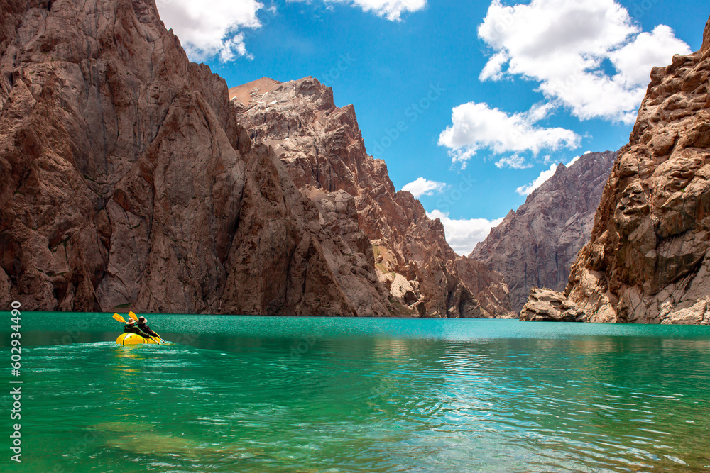 Obraz premium Kayaking on a mountain lake. Two men are sailing on a red canoe along the lake along the rocks. The theme of water sports and summer holidays.