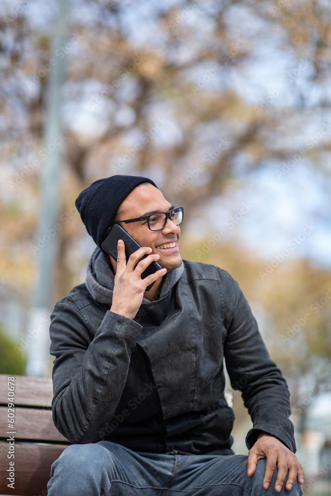 Smiling young african man in winter wear sitting on bench outside talking on phone