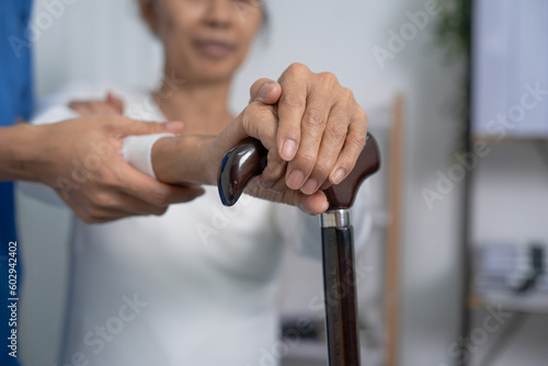 Attentive practitioner nurse assisting physical therapy elderly woman on a walking wood standard cane in disability nursing rehabilitation center, physical therapy encourage hands.