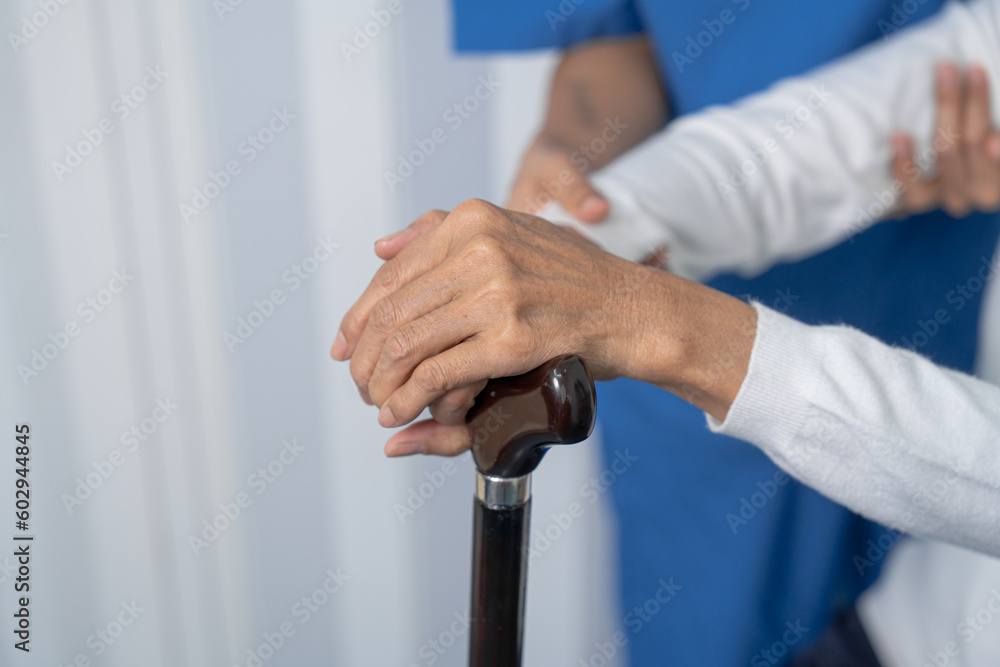Attentive practitioner nurse assisting physical therapy elderly woman on a walking wood standard cane in disability nursing rehabilitation center, physical therapy encourage hands.