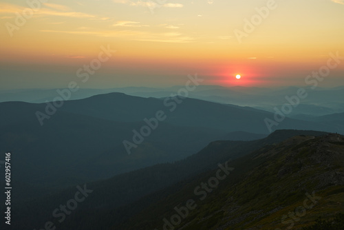 Sunrise in mountains. Natural mountain landscape with illuminated misty peaks  foggy slopes and valleys  blue sky with orange yellow sunlight