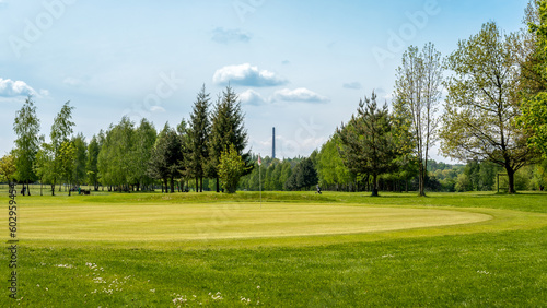 Golf course located in Bazantarnia Park in Siemianowice, Silesia, Poland. Perfectly cutted lawn surrounded by fresh trees. Fresh, awakening nature in May. Industrial chimney in the background.