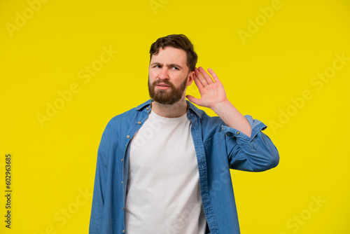 Young, serious, blond man in a blue shirt on a yellow background, holding an ear to listen better, what you're saying. Photo Shoot.