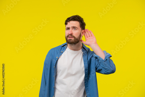 Quadro su tela Young, serious, blond man in a blue shirt on a yellow background, holding an ear to listen better, what you're saying