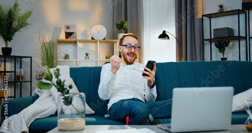 Attractive happy in high spirits young bearded man in glasses rejoycing when get good news on smartphone and raising his hands from happiness