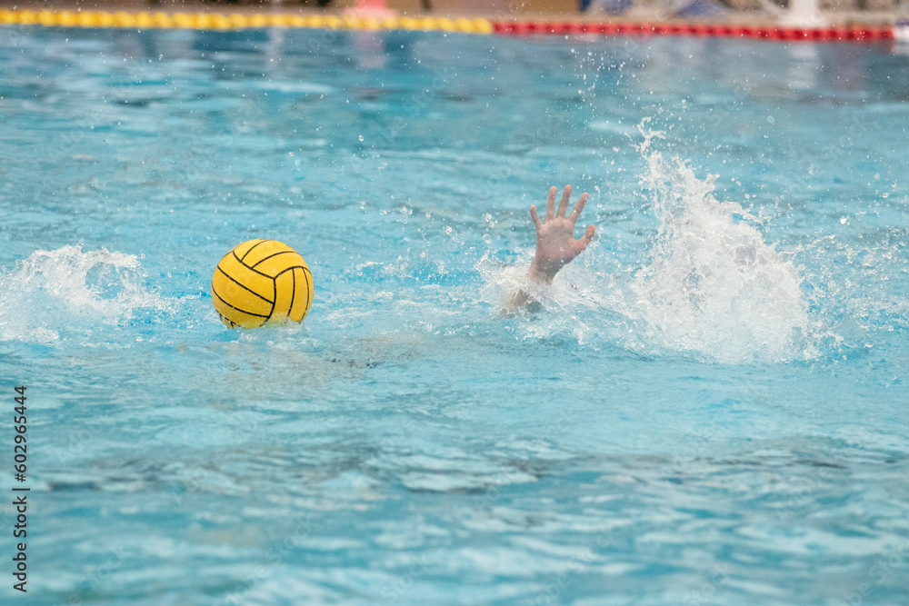 One hand of a Caucasian water polo player reaching up through the surface of the water in the swimming pool to grab the yellow water polo ball floating on the water's surface.