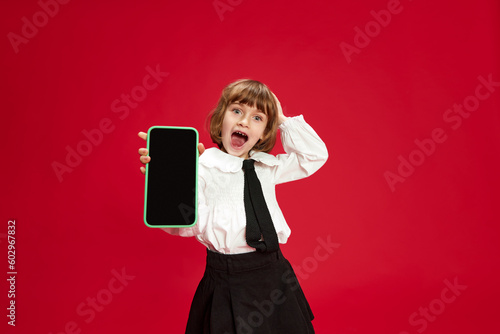 Portrait of happy, smiling, laughing little girl in white blouse and black shirt, showing mobile phone screen against red studio background. Concept of childhood, education, fashion, kid emotions photo