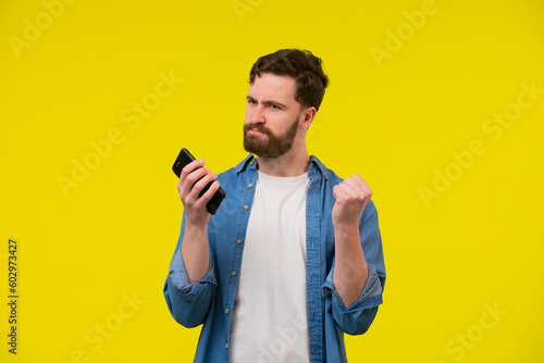 Overjoyed happy tanned handsome man in blue basic t-shirt raise fist up using phone posing isolated on orange yellow studio background. Copy space Banner Mockup. Online Social media. Lucky win concept