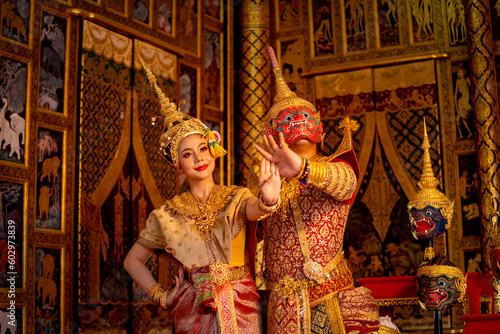 Thai classic masked man from the Ramakien character with red mask dance with beautiful Asian woman wear Thai traditional dress and stay in front of Thai painting on public place wall. photo