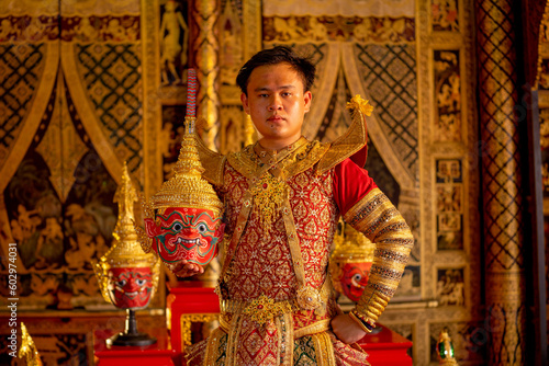 Portrait of Asian man hold Pantomime or Khon masks with red giant character and look at camera also stay in front of Thai painting on wall in public place.